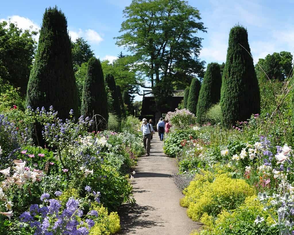 Een weelderig tuinpad omzoomd door kleurrijke bloeiende bloemen en hoge, smalle groenblijvende bomen, met een paar mensen die wandelen en genieten van de zonnige dag.