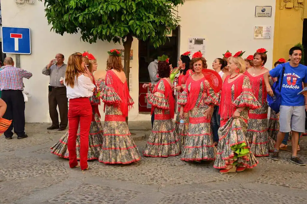 Een groep vrouwen in kleurrijke traditionele flamencojurken en rode sjaals komt samen en praat op een geplaveide straat, met een aantal mensen in de buurt die toekijken en een boom op de achtergrond.