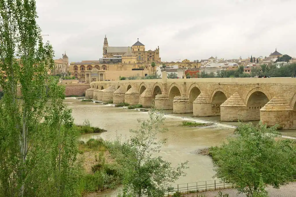 Een historische stenen brug steekt een brede rivier over met groen langs de oevers. Op de achtergrond steken een grote kathedraal en andere oude gebouwen uit boven de stad Córdoba, Spanje, onder een bewolkte hemel.