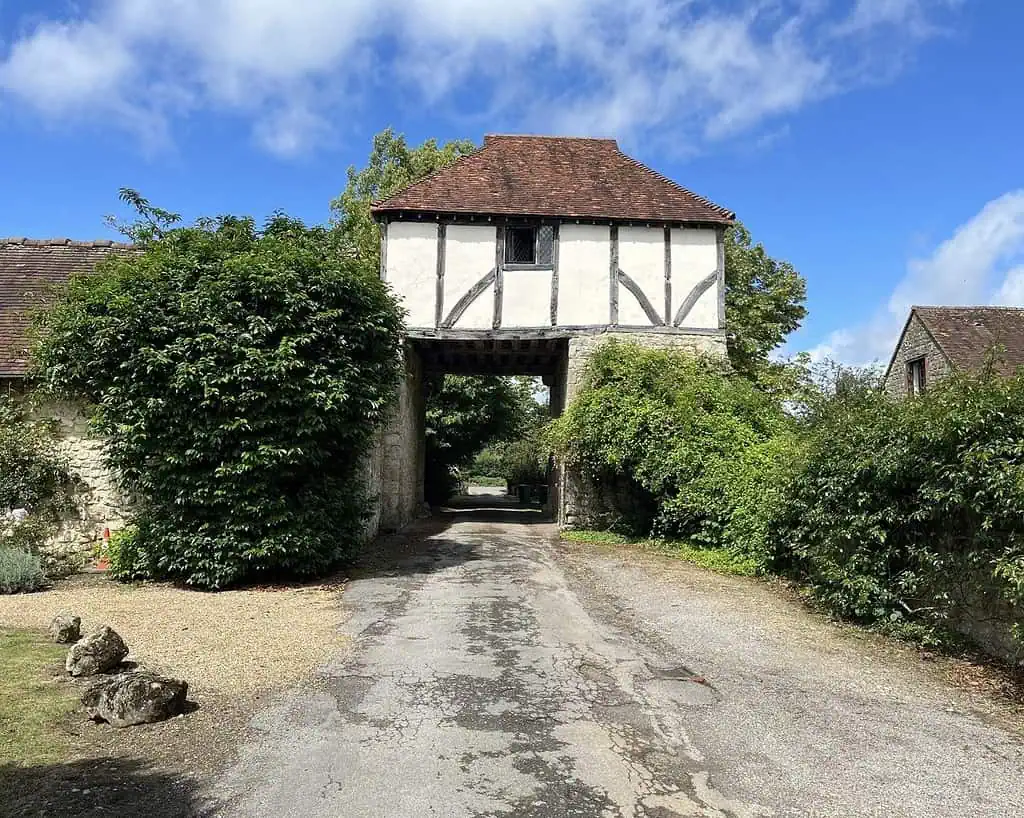 Een smal landweggetje in Oxfordshire loopt onder een vakwerkpoort met een rood pannendak door, omringd door groene struiken en bomen, onder een helderblauwe lucht met wolken.