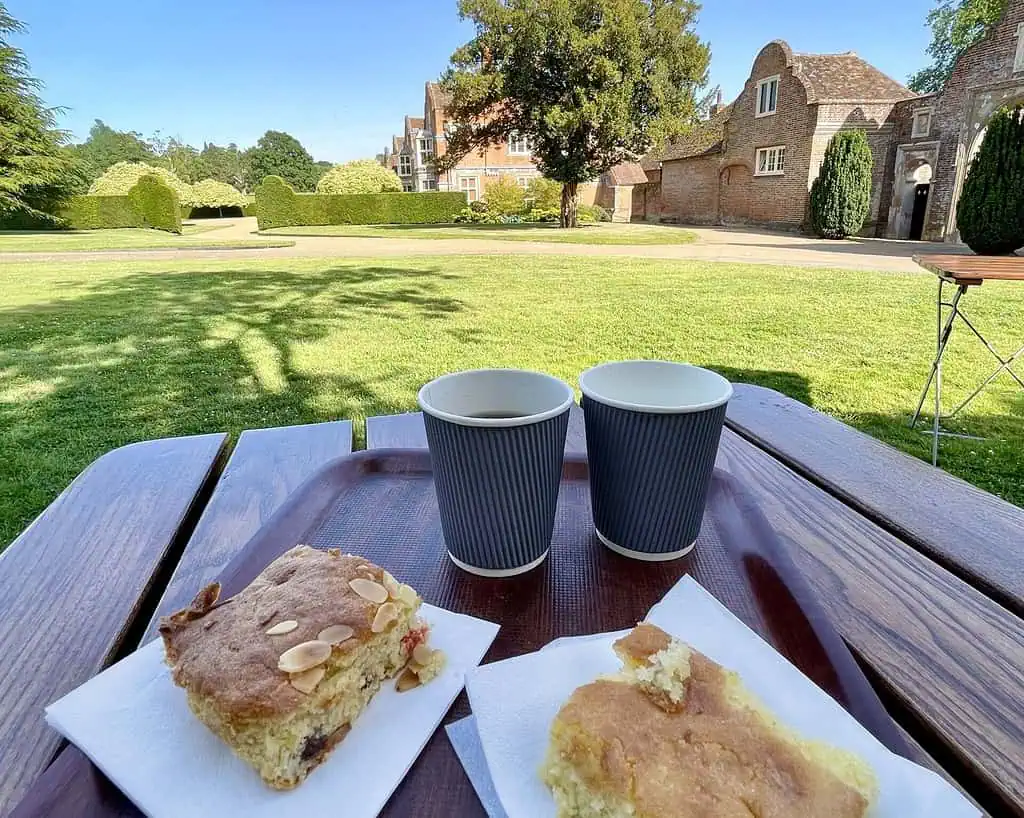 Twee papieren kopjes koffie en twee stukken cake rusten op servetten op een houten picknicktafel, tegen een zonnig gazon in Oxfordshire met oude bakstenen gebouwen en bomen op de achtergrond.