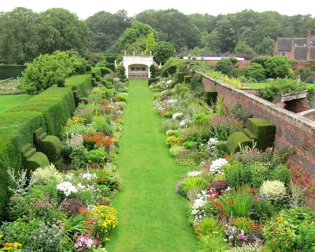 Een goed onderhouden tuin met een centraal groen gazon, omzoomd door kleurrijke bloembedden en netjes gesnoeide heggen, met bakstenen muren en bomen op de achtergrond.