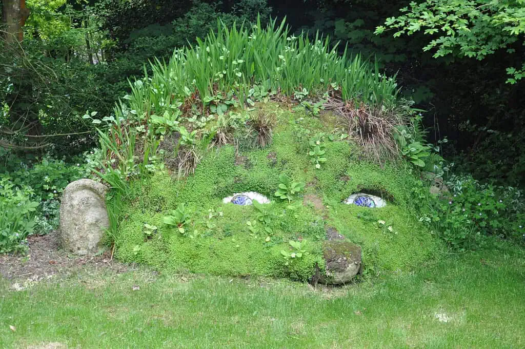 Een grasheuvel in de vorm van een gezicht met blauwe en witte ogen, neus en mond, bedekt met mos en planten, ligt in een Cornwall-tuin omringd door bomen en weelderig groen - een betoverend gezicht op elke voorjaarsreis.