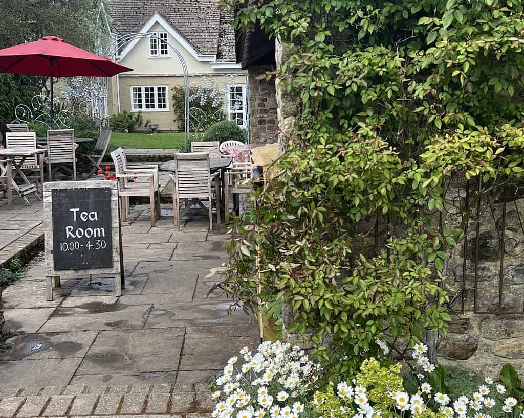 Buitenpatio met houten tafels en stoelen, een rode paraplu en een bord met daarop Tea Room 10:00 - 4:30 uur. Bloemen en groen omringen het gebied, met een charmante Cornwall cottage in het zicht - perfect voor je voorjaarsreis.