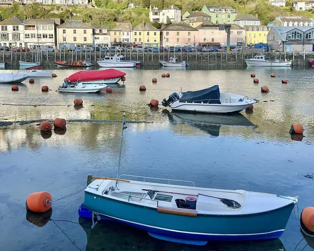 Kleine bootjes dobberen op kalm water in een haven in Cornwall, omringd door oranje boeien. Kleurrijke huizen en gebouwen omzomen de heuvels op de achtergrond onder zacht daglicht, waarmee de charme van een voorjaarsreis wordt vastgelegd.