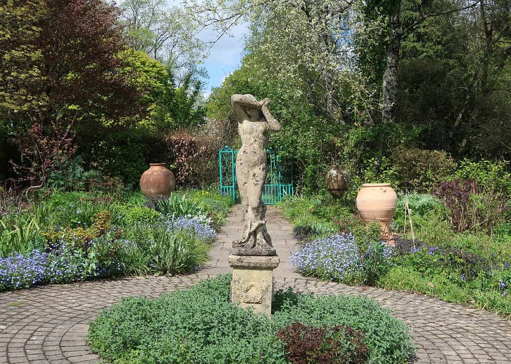Een verweerd stenen beeld zonder hoofd staat op een sokkel in het midden van een cirkelvormig tuinpad in Cornwall, omringd door groen, bloeiende bloemen, grote potten van klei en een groen metalen hek op de achtergrond.