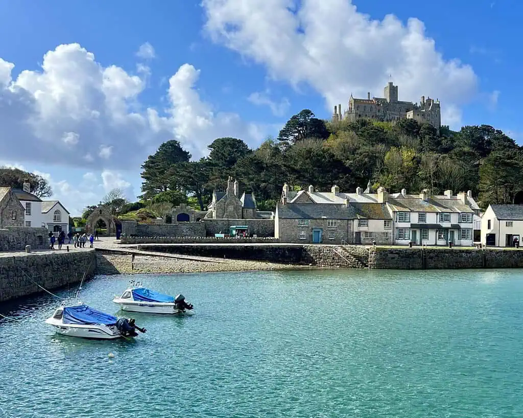 Kleine bootjes dobberen in een turquoise haven in Cornwall in de buurt van stenen huisjes, met een historisch kasteel hoog op een heuvel omringd door bomen onder een blauwe hemel. Mensen op hun voorjaarsreis wandelen langs de schilderachtige waterkant.
