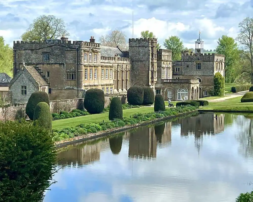 Een groot historisch stenen herenhuis met torentjes en boogramen staat naast een rustige spiegelende gracht in Cornwall, omringd door verzorgde tuinen, vormsnoeibossen en weelderige groene bomen onder een gedeeltelijk bewolkte hemel - perfect voor een voorjaarsreis.