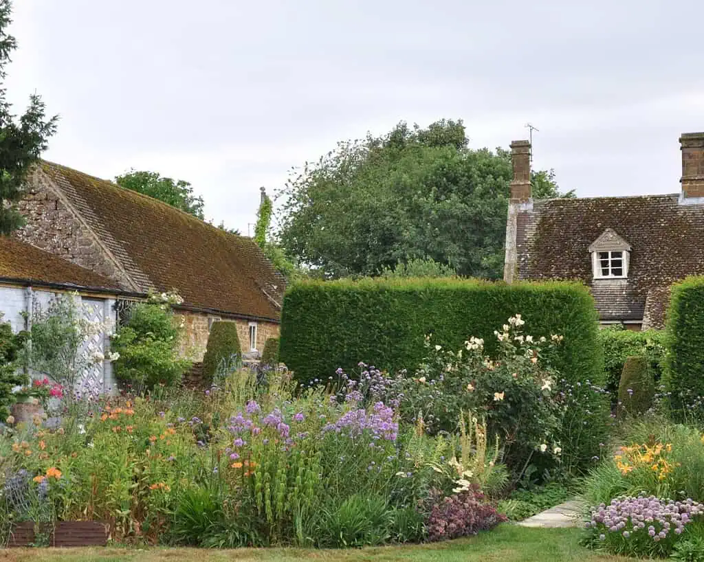 Een cottage tuin met een mix van kleurrijke bloemen en weelderig groen, omzoomd door hoge heggen. Op de achtergrond staan rustieke stenen gebouwen met bemoste, schuine daken en schoorstenen onder een bewolkte hemel.