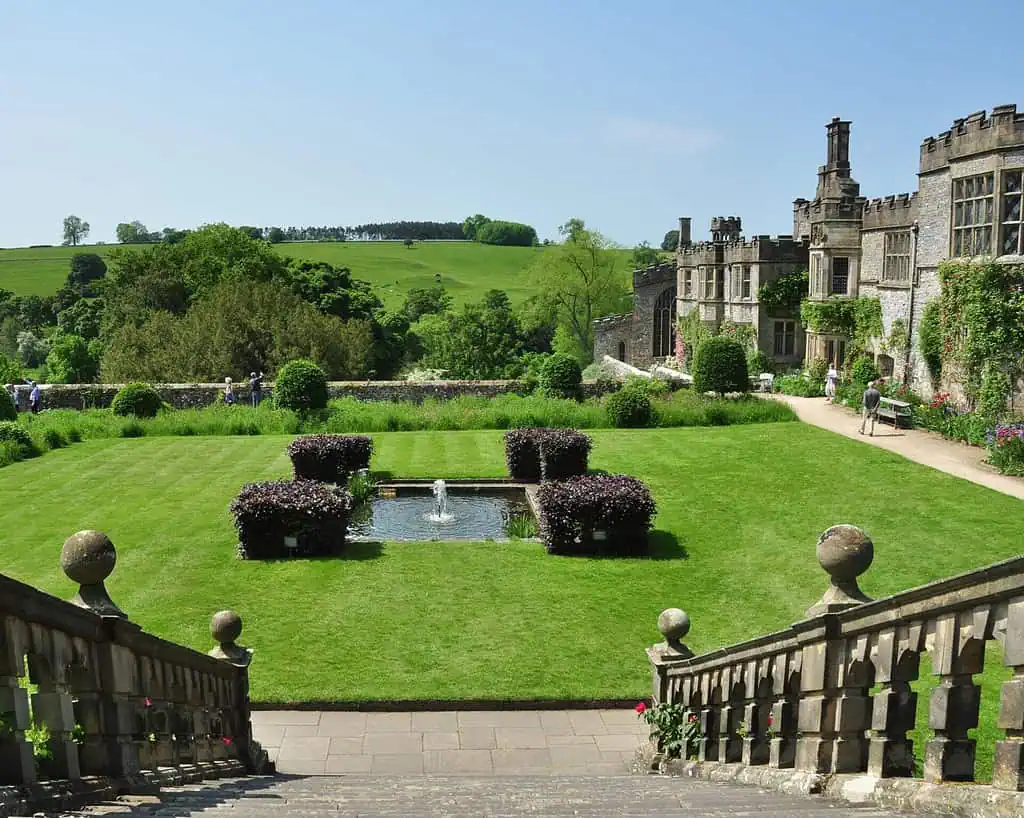 Stenen trappen leiden naar een gemanicuurd gazon met een kleine centrale fontein, netjes gesnoeide struiken en een sierlijk historisch gebouw aan de rechterkant - perfect om het rustig aan te doen in de glooiende groene heuvels van Derbyshire onder een helderblauwe hemel.