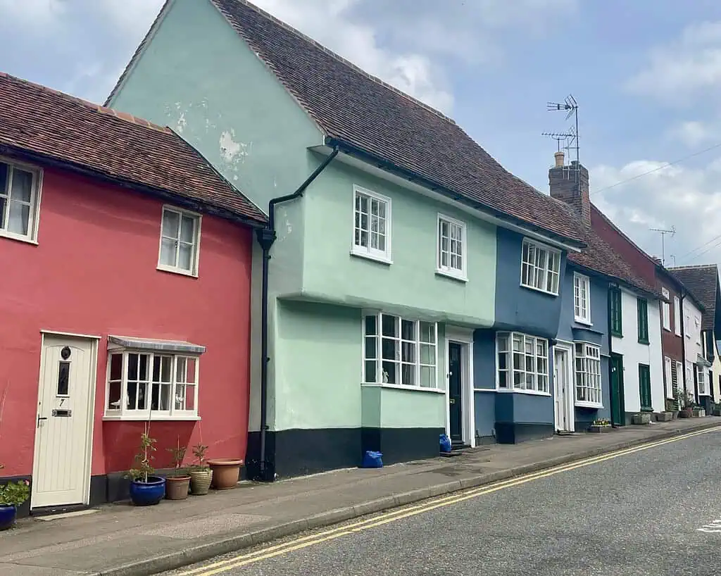Een rij kleurrijke huizen met rode, groene, blauwe en witte gevels omzoomt een rustige straat in Derbyshire. Verschillende potplanten staan bij de deuren en de halfbewolkte lucht draagt bij aan de sfeer van Taking it Easy.