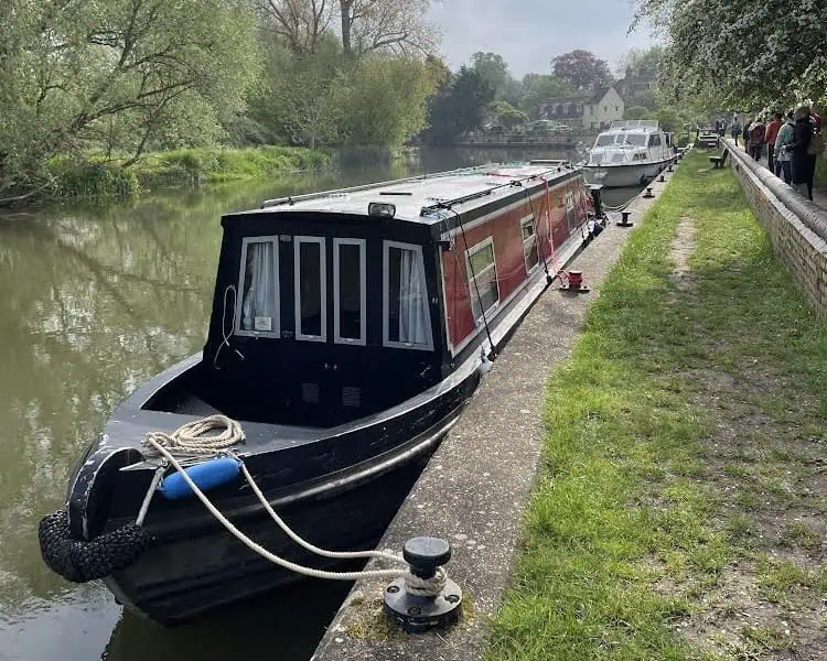 Een narrowboat met een rode en zwarte buitenkant ligt aangemeerd langs een rustig kanaal in Derbyshire, met bomen aan de ene kant en een stenen pad waar mensen het rustig aan doen. Op de achtergrond zijn nog een boot en gebouwen te zien.