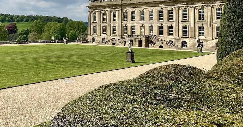 Een groots historisch herenhuis met sierlijke stenen architectuur staat naast een groot gemanicuurd gazon en grindpaden in Derbyshire, omringd door bomen onder een gedeeltelijk bewolkte hemel - perfect om het rustig aan te doen.