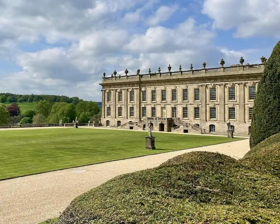 Een groots historisch herenhuis met sierlijke stenen architectuur staat naast een groot gemanicuurd gazon en grindpaden in Derbyshire, omringd door bomen onder een gedeeltelijk bewolkte hemel - perfect om het rustig aan te doen.