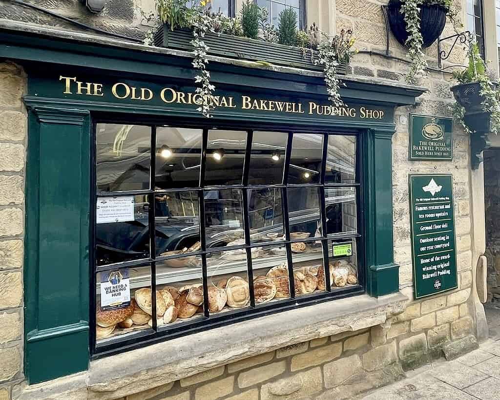 Een bakkerijetalage in The Old Original Bakewell Pudding Shop in Derbyshire toont rijen broodbroden en gebak. De winkel heeft groene versieringen, hangende bloemen en stenen muren met borden aan de buitenkant, perfect om het rustig aan te doen.