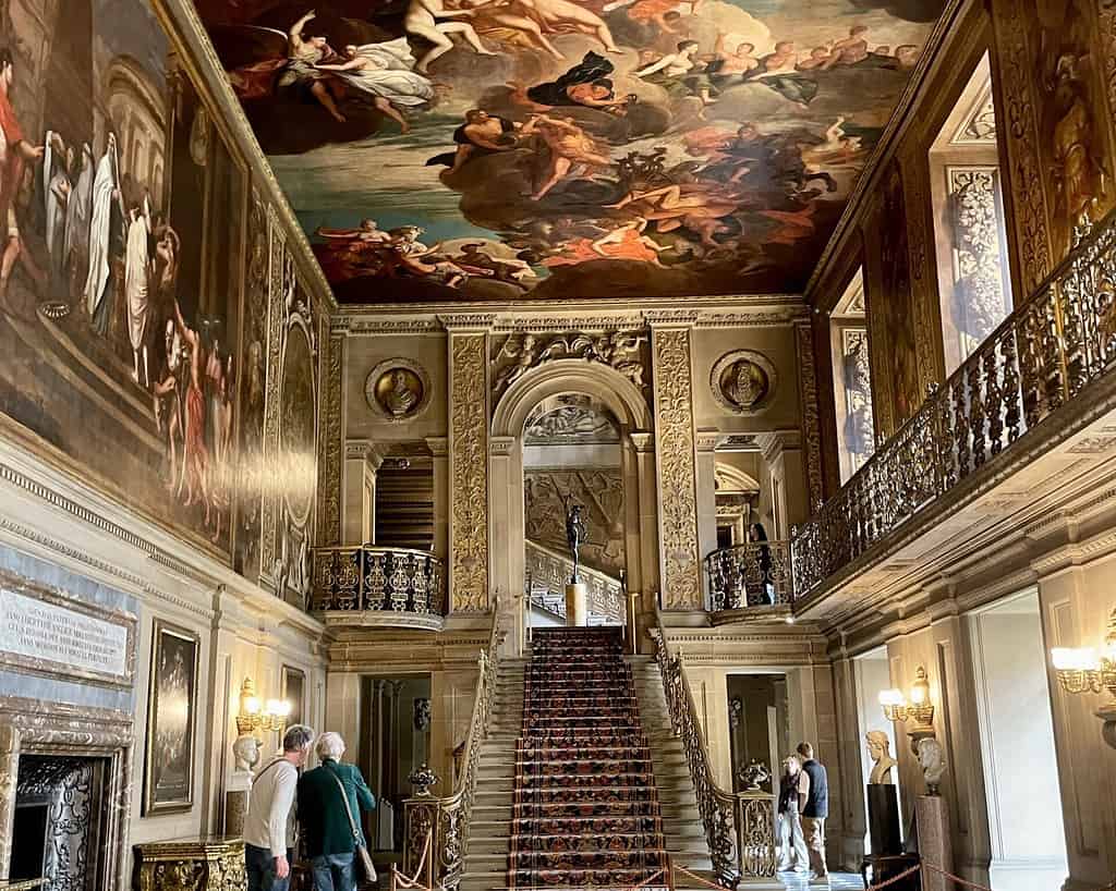 Een grote, sierlijke trap in Derbyshire met een rood tapijt leidt naar een gewelfde deuropening, omringd door marmeren beelden, gouden balustrades en grote klassieke schilderijen. Verschillende mensen doen het rustig aan terwijl ze de weelderige hal bewonderen.