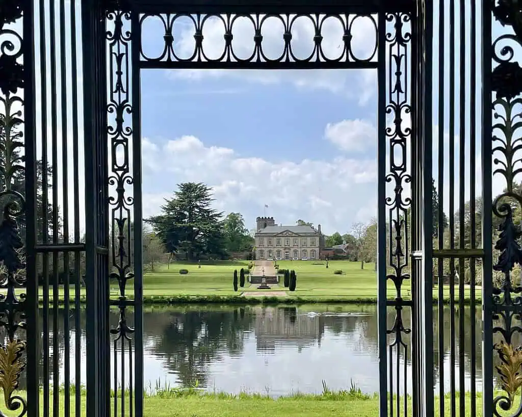 Een groot landhuis in Derbyshire ligt aan de overkant van een weerspiegelende vijver, met uitzicht door sierlijke ijzeren hekken, met verzorgde groene gazons en bomen onder een heldere, halfbewolkte hemel - perfect om het rustig aan te doen.
