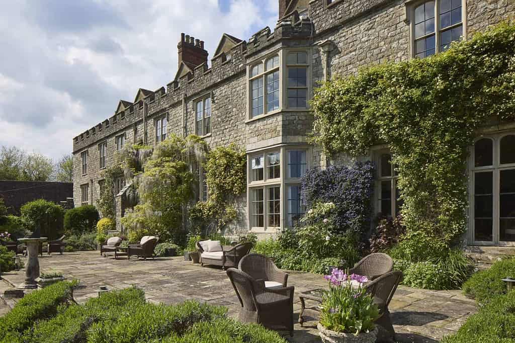 Een historisch stenen landhuis met klimplanten, grote ramen en een patio met rieten stoelen en tafels. Weelderige tuinen en bloemen in potten omringen de zonnige zithoek buiten.