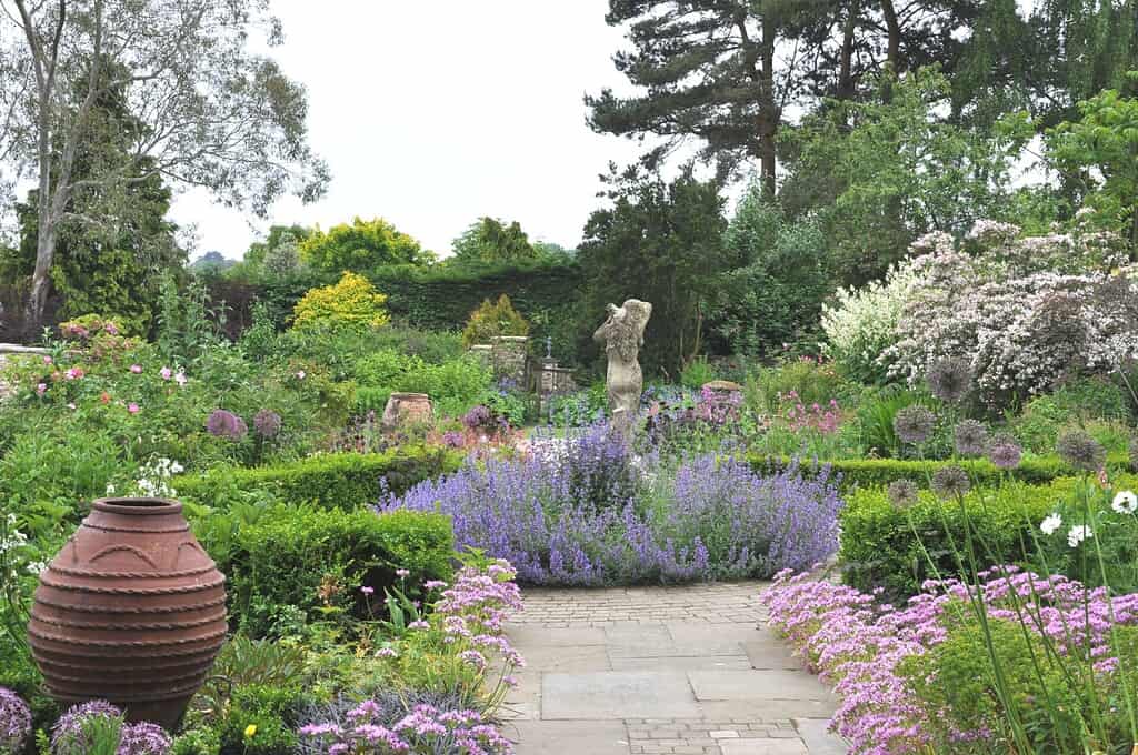 Een weelderige tuin met een stenen pad, levendige paarse, roze en witte bloemen, groene struiken, een urn aan de linkerkant en een centraal standbeeld omringd door bloeiende planten en hoge bomen op de achtergrond.