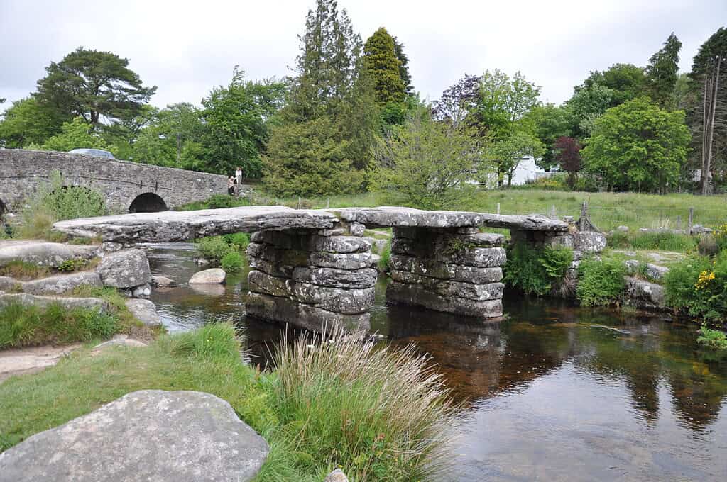 Een oude stenen klapbrug met grote vlakke platen rust op stenen pilaren over een helder beekje, omringd door gras, bomen en een stenen boogbrug op de achtergrond.