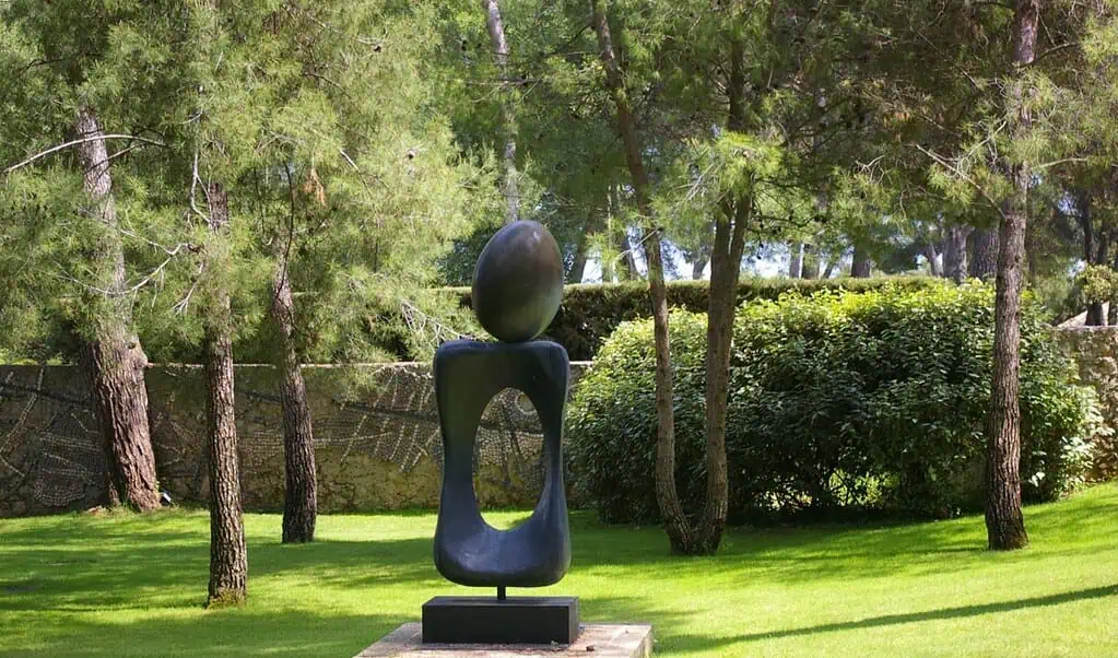 Een moderne abstracte sculptuur met een gladde ovale vorm gebalanceerd op een holle, gebogen sokkel staat op een sokkel in de zonovergoten tuin van Fondation Maeght, omringd door bomen en weelderig groen.