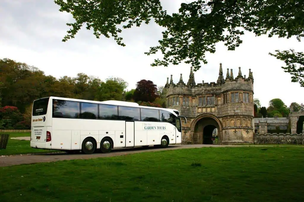 Een witte tourbus met het label Garden Tours staat geparkeerd op een grasveld voor een historisch stenen poortgebouw met gewelfde ingangen en sierlijke architectuur, omringd door bomen. Neem contact op voor meer informatie over de tour.