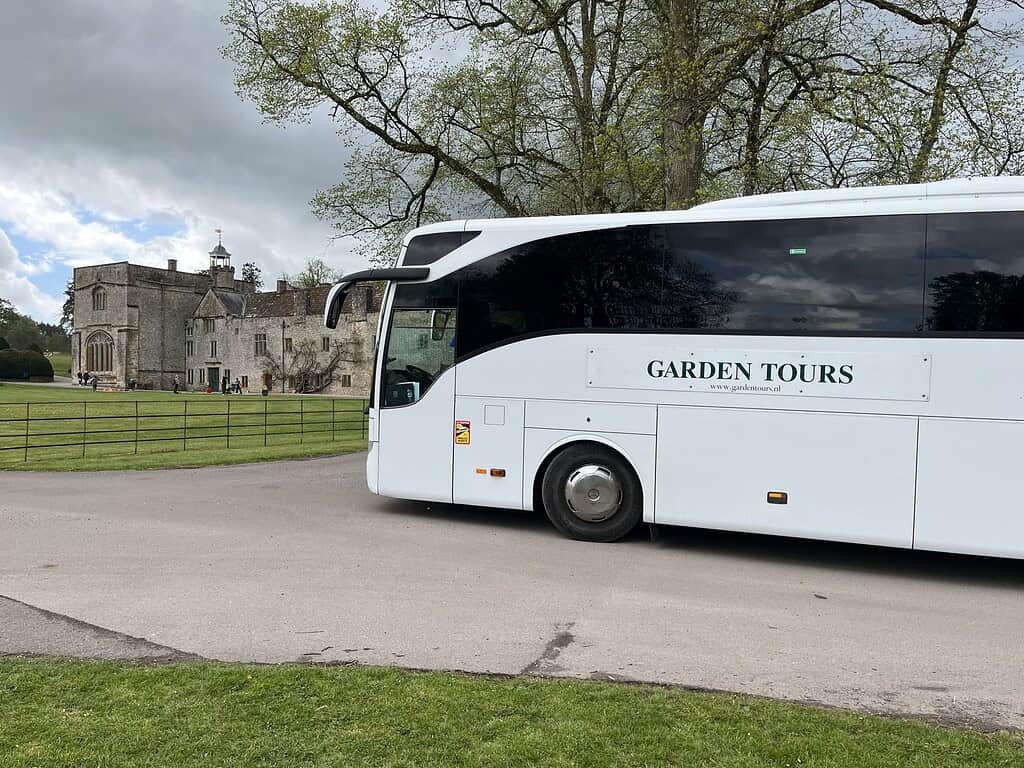 Een witte tourbus met de naam Garden Tours staat geparkeerd op een verharde weg bij een groot historisch stenen gebouw omringd door groene gazons en bomen onder een bewolkte hemel.