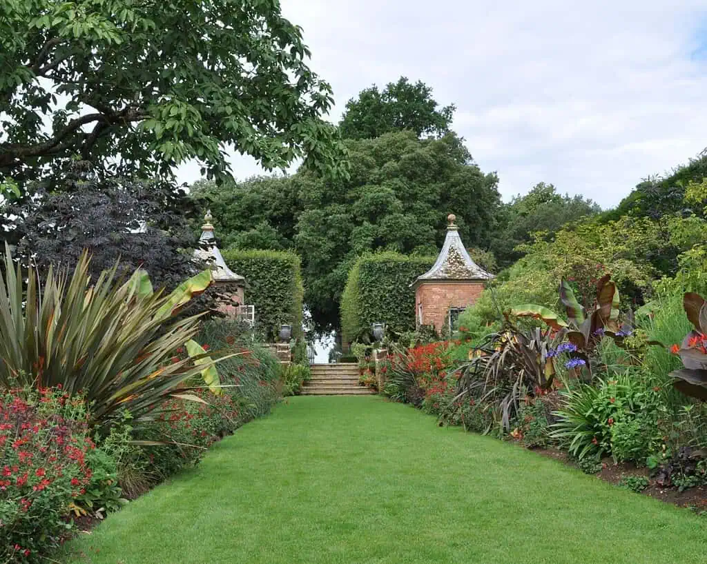 Een weelderig groen tuinpad met kleurrijke bloemen en planten leidt naar stenen trappen geflankeerd door twee kleine, sierlijke bakstenen paviljoens, met hoge bomen en heggen op de achtergrond onder een gedeeltelijk bewolkte hemel.