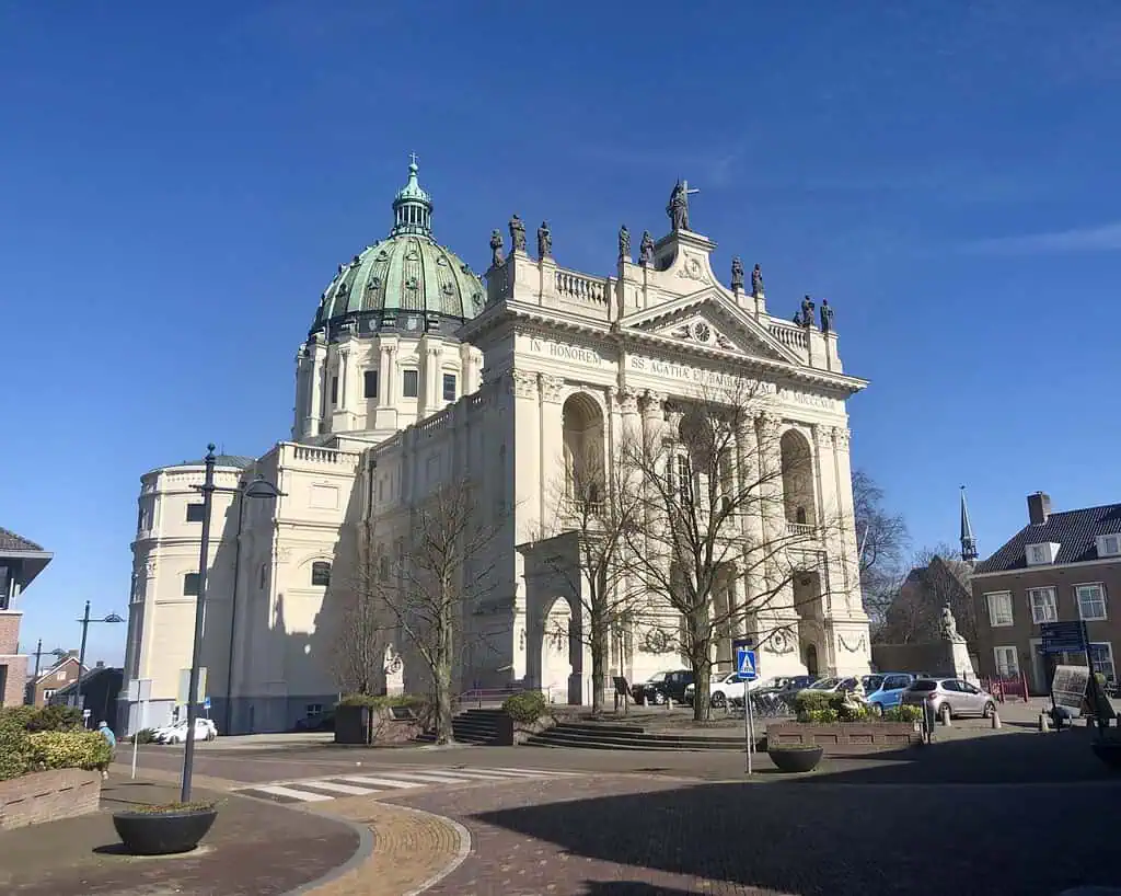 Een grote, historische, witte kerk met een groene koepel en beelden op het dak staat onder een strakblauwe hemel, omringd door bloeiende bomen en geparkeerde auto's, die de essentie van Nederland in bloei op een zonnige dag vastlegt.