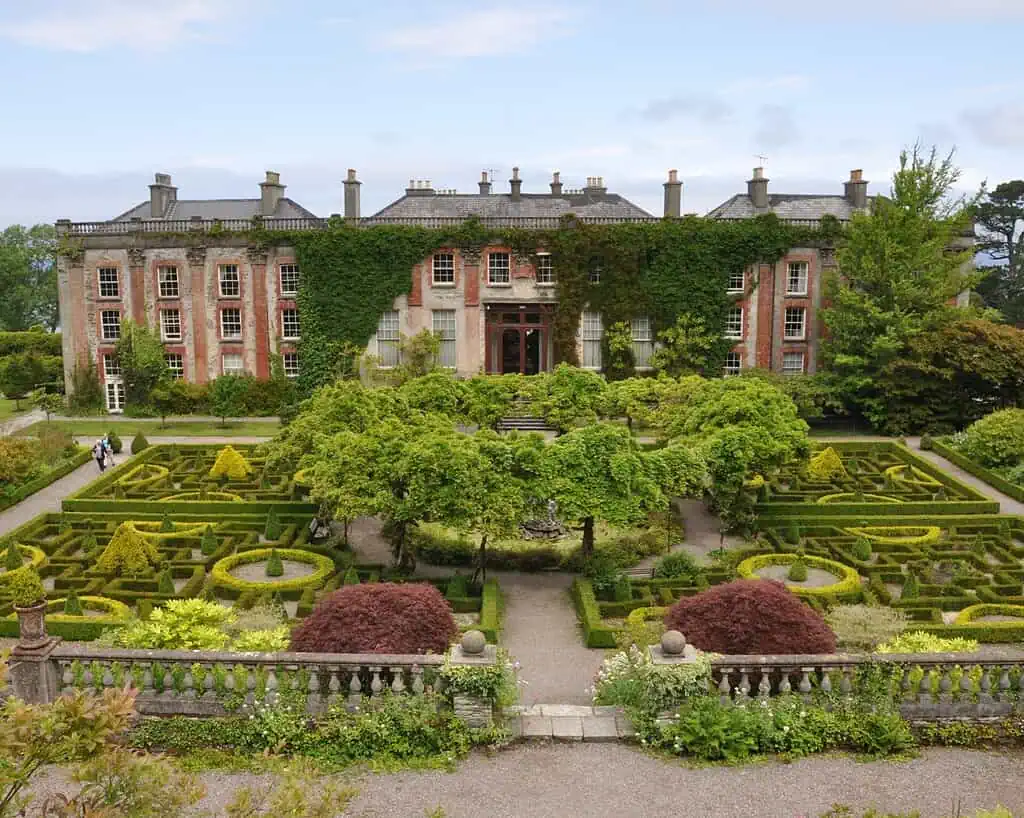 Een groot historisch herenhuis met met klimop bedekte muren staat achter een sierlijke formele tuin in Ierland, met symmetrische heggen, grindpaden, gevormde struiken en weelderig groen met groene accenten onder een gedeeltelijk bewolkte hemel.