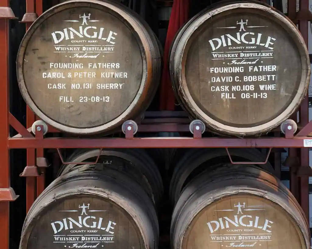Vier houten whiskeyvaten met groene accenten staan in twee rijen op elkaar. Elk vat is gemerkt met Dingle Whiskey Distillery, inclusief de gegevens van de oprichters, het nummer van het vat, de afvuldatum en het whiskytype uit Ierland.