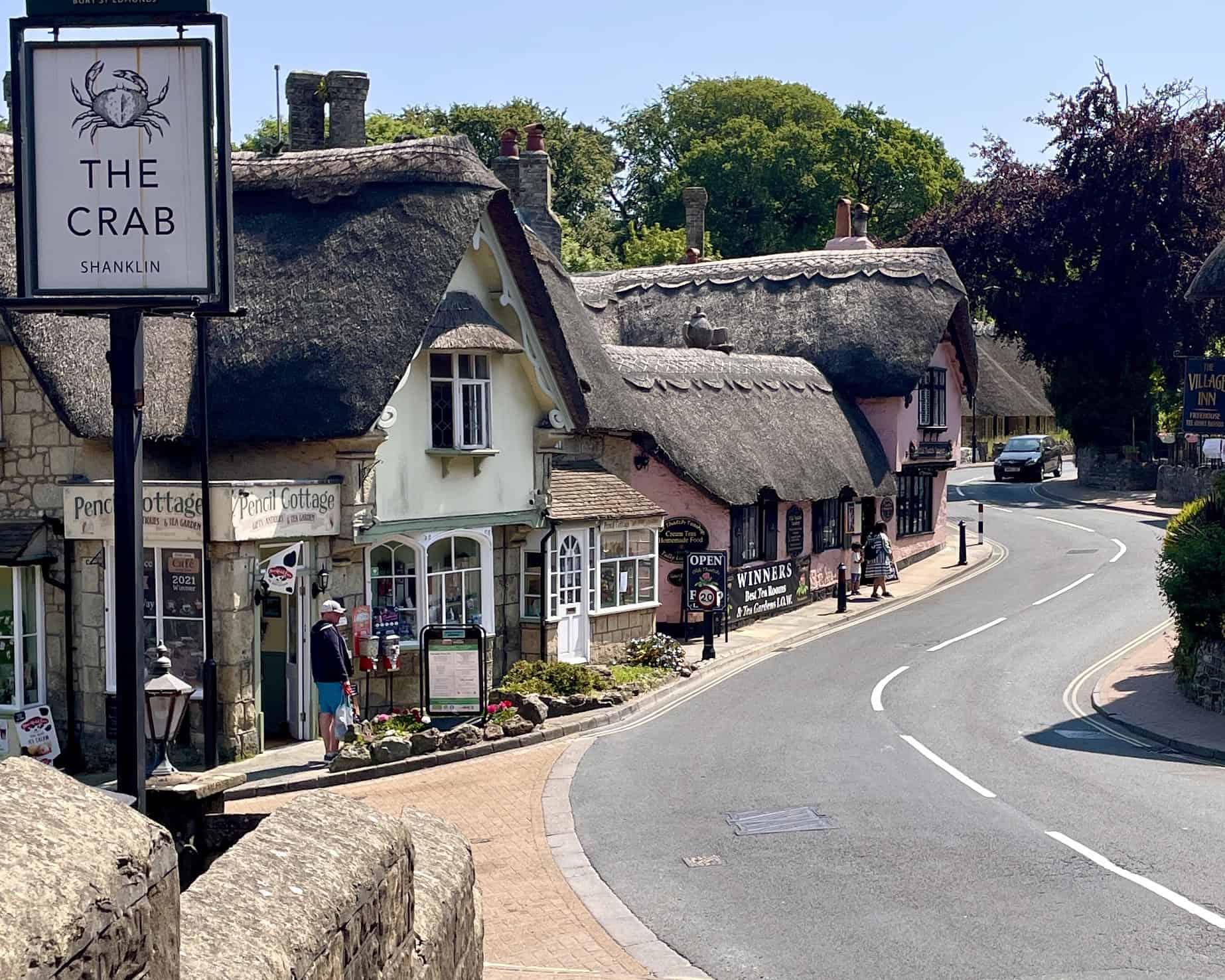 Een gebogen straat in Shanklin op het Isle of Wight met charmante huisjes met rieten daken, winkels en restaurant The Crab. Mensen wandelen op het trottoir op een zonnige dag met groene bomen op de achtergrond.