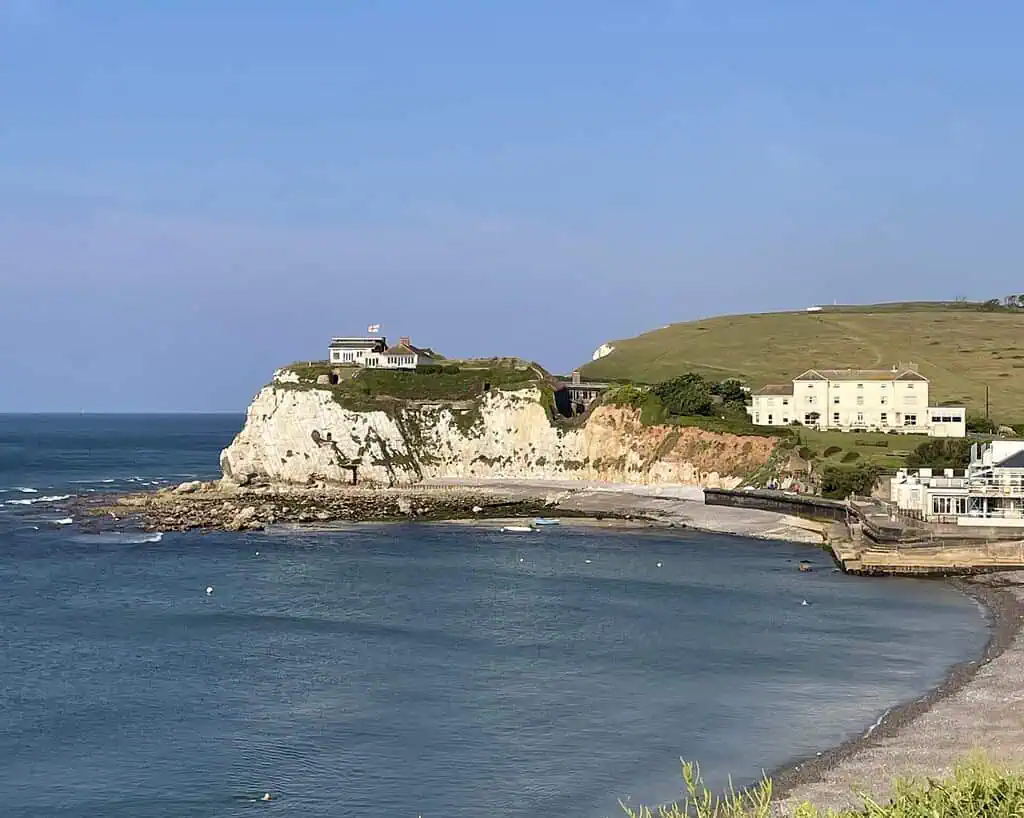 Een kusttafereel op het Isle of Wight met witte krijtrotsen, een huis met een vlag erop, met gras begroeide heuvels en verschillende gebouwen met uitzicht op een kalme blauwe zee en een klein, gebogen strand onder een heldere hemel.