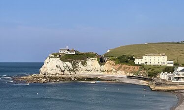 Een kusttafereel op het Isle of Wight met witte krijtrotsen, een huis met een vlag erop, met gras begroeide heuvels en verschillende gebouwen met uitzicht op een kalme blauwe zee en een klein, gebogen strand onder een heldere hemel.