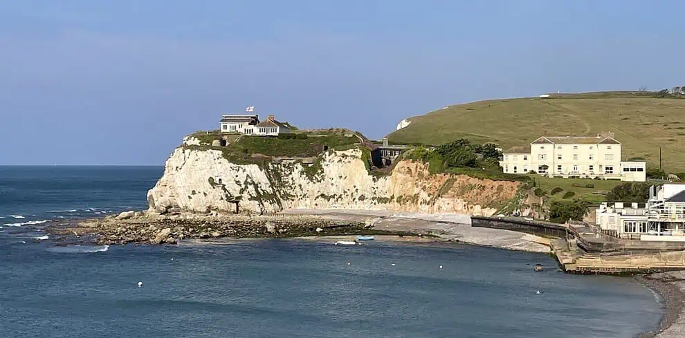 Een kusttafereel op het Isle of Wight met witte krijtrotsen, een huis met een vlag erop, met gras begroeide heuvels en verschillende gebouwen met uitzicht op een kalme blauwe zee en een klein, gebogen strand onder een heldere hemel.