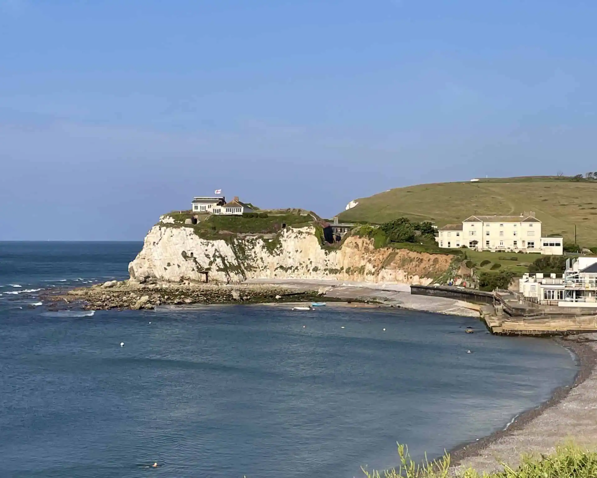 Een kusttafereel op het Isle of Wight met witte krijtrotsen, een huis met een vlag erop, met gras begroeide heuvels en verschillende gebouwen met uitzicht op een kalme blauwe zee en een klein, gebogen strand onder een heldere hemel.
