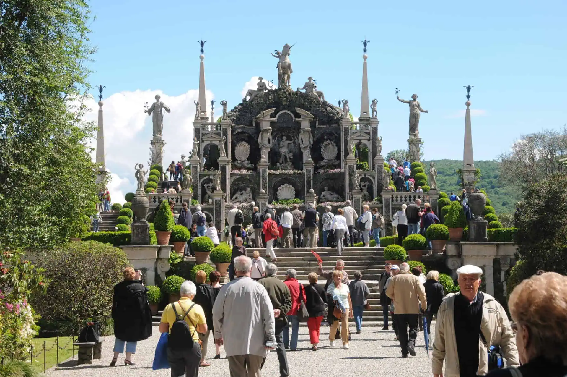 Een groep toeristen loopt naar een sierlijk barok tuinterras met beelden, zuilen en hagen op Isola Bella onder een strakblauwe hemel.