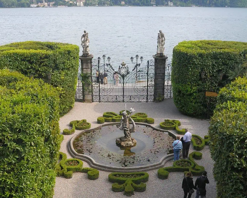 Een formele tuin met een ronde fontein en een standbeeld in het midden, omringd door verzorgde heggen. Vier mensen staan in de buurt. Sierlijke poorten en standbeelden kijken uit over een groot meer, dat doet denken aan de elegantie van Italiaanse meren, met gebouwen aan de verre oever.