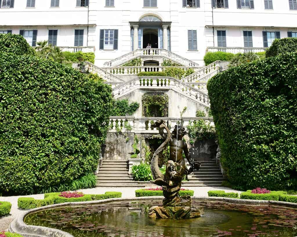 Een grote trap met sierlijke leuningen leidt naar een wit gebouw met blauwe luiken, dat doet denken aan elegante villa's aan de Italiaanse meren, omringd door weelderige groene hagen en een fontein met een standbeeld dat een grote vis vasthoudt.