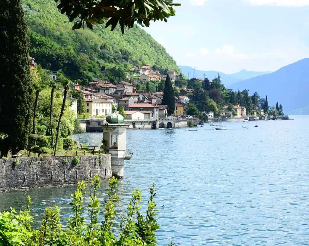 Een schilderachtig dorp aan het meer met huizen genesteld op een groene heuvel, bomen en een kleine structuur aan het water naast kalm blauw water, dat doet denken aan de serene schoonheid rond de Italiaanse meren, met bergen zichtbaar op de achtergrond.