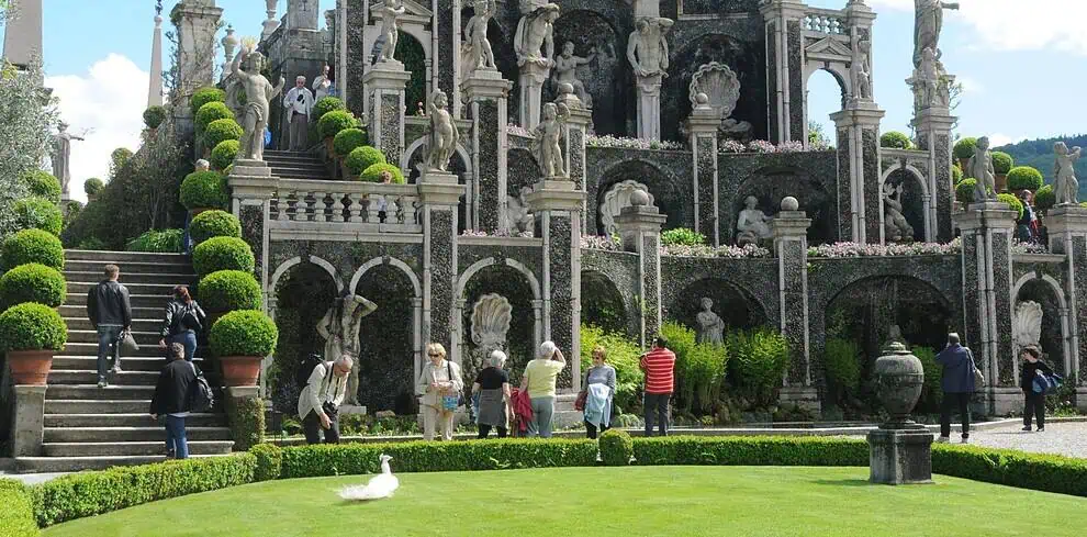 Bezoekers verkennen een sierlijke terrastuin die is geïnspireerd op de elegantie van de Italiaanse meren, met grote stenen beelden, bogen en keurig gesnoeide vormsnoei. Een groot groen gazon ligt op de voorgrond onder een blauwe lucht met wolken.