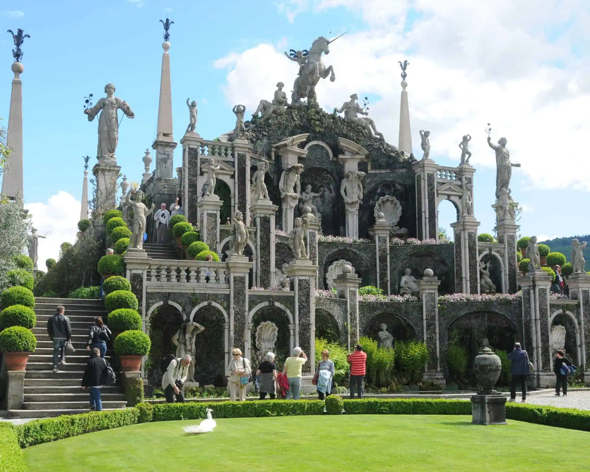 Bezoekers verkennen een sierlijke terrastuin die is geïnspireerd op de elegantie van de Italiaanse meren, met grote stenen beelden, bogen en keurig gesnoeide vormsnoei. Een groot groen gazon ligt op de voorgrond onder een blauwe lucht met wolken.