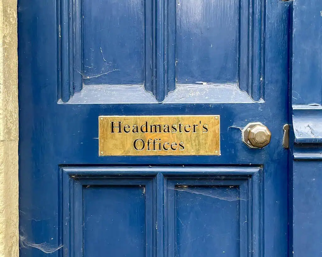 Een blauwe houten deur met een koperen bordje met de tekst Headmasters Offices en een ronde metalen deurknop aan de rechterkant, die doet denken aan het soort ingang dat je zou kunnen vinden als je Jane Austens voetsporen volgt op literaire reizen.