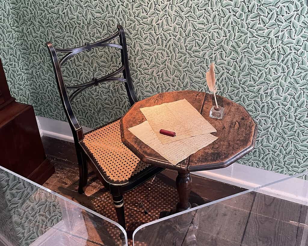 Op een kleine houten tafel, die doet denken aan de tijd van Jane Austen, liggen handgeschreven brieven, een rode zegellak en een ganzenveer in een inktpot naast een geweven stoel tegen een behang met groene bladmotieven.