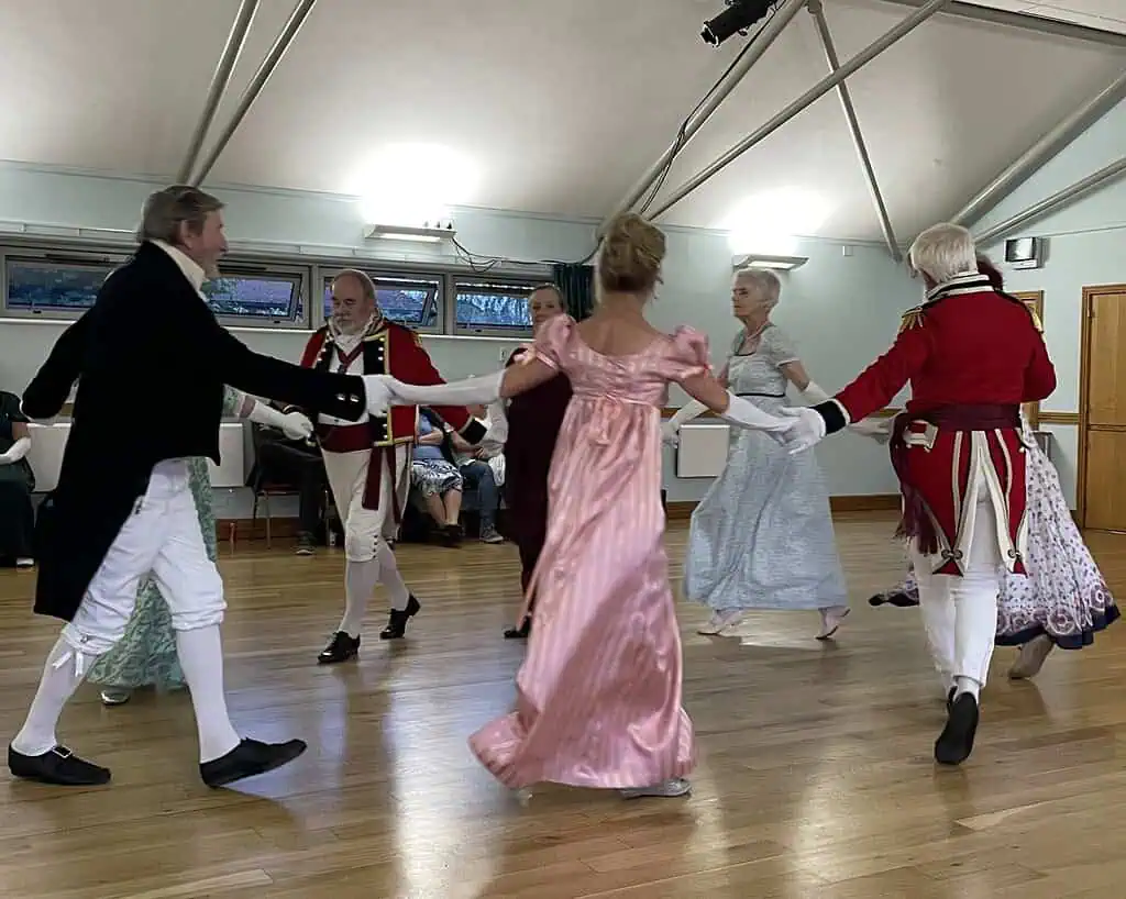 Een groep volwassenen in kostuums uit de tijd van Jane Austen danst in een cirkel in een helder verlichte zaal. De mannen dragen jacquet of militaire uniformen en de vrouwen elegante jurken. Anderen zitten en kijken toe op de achtergrond.