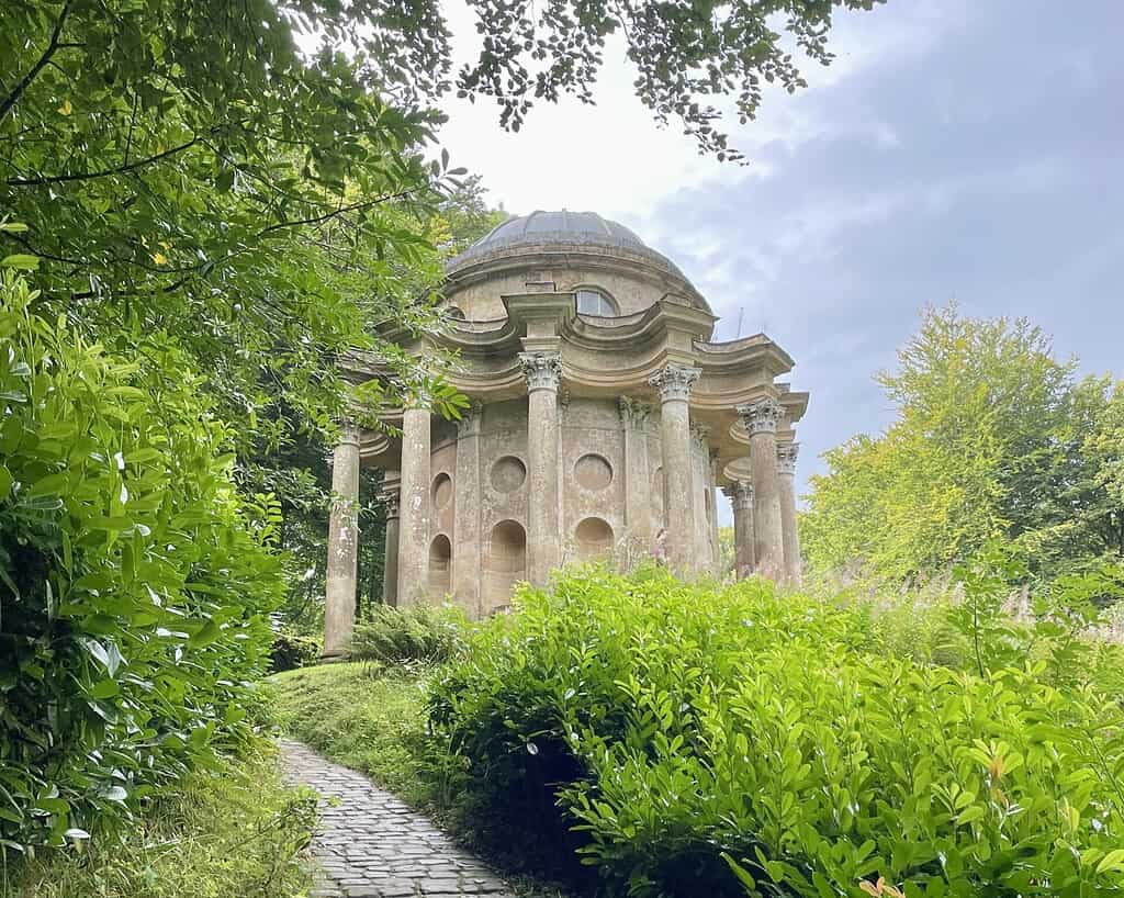 Een stenen pad leidt door weelderig groen struikgewas naar een historisch, koepelvormig stenen paviljoen met hoge zuilen en ronde ramen, dat doet denken aan een Jane Austen-setting, omringd door bomen onder een gedeeltelijk bewolkte hemel.