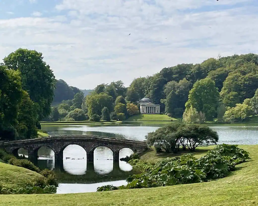 Een stenen brug bogen over een rustig meer omringd door weelderige groene bomen en gras, echo's van de tijdloze elegantie van een Jane Austen reis, met een klassieke paviljoen zichtbaar in de verte onder een gedeeltelijk bewolkte hemel.