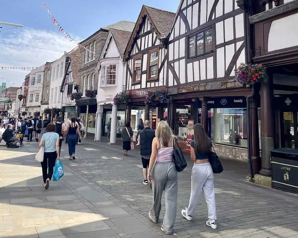 Mensen lopen over een geplaveide voetgangersstraat met vakwerkhuizen en winkels in historische stijl, in de voetsporen van Jane Austen. Hangende bloemenmanden en feestelijke slingers sieren het tafereel onder een blauwe hemel.