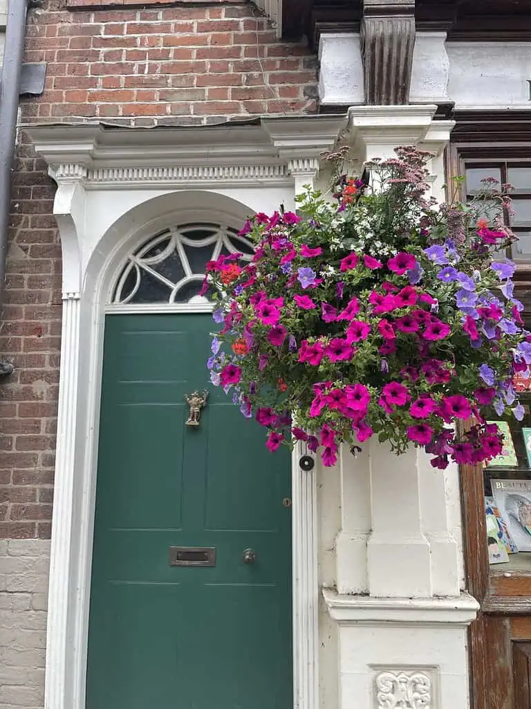 Een groene deur met een koperen klopper wordt omlijst door een boog, die doet denken aan de tijd van Jane Austen. Aan de rechterkant overstroomt een grote hangende mand met levendige roze en paarse bloemen tegen de rode bakstenen - een tafereel dat past bij Jane Austen reizen.