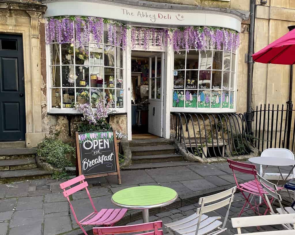 Een charmante delicatessenzaak met een gebogen raam, gedrapeerd in paarse blauwe regen, doet denken aan een scène uit Jane Austen. Het bord OPEN voor Ontbijt verwelkomt je in kleurrijke stoelen en tafels die buiten op de stenen stoep staan.